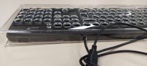 Foto colorida aproximada de um teclado padrão de computador preto, com cabo USB, e com uma colmeia em acrílico transparente encaixada nele.