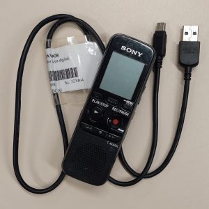 Foto colorida de gravador de voz preto da marca Sony, que está sobre seu cabo carregador USB..