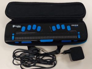 Foto colorida de uma linha Braille dentro de um estojo aberto. O interior do estojo e o equipamento são pretos e os botões do equipamento são azuis. O cabo do conexão e a fonte de carregamento estão em frente ao estojo.