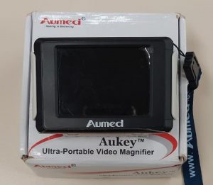 Foto colorida de uma lupa eletrônica da marca Aumed sobre a caixa. A lupa é preta e está presa a uma alça de tecido azul. E a caixa é branca com letras escritas em preto e vermelho.