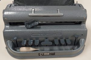 Foto colorida de uma máquina Braille da marca Perkins cinza, com as teclas cinzas um pouco mais escuras. Junto com ela há um apagador em madeira.